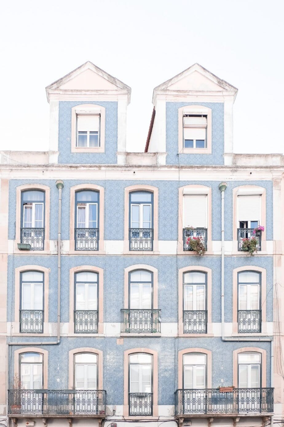 A four-story light blue building with white trim, iron balconies, and two dormer windows on the roof. Some balconies have potted plants.