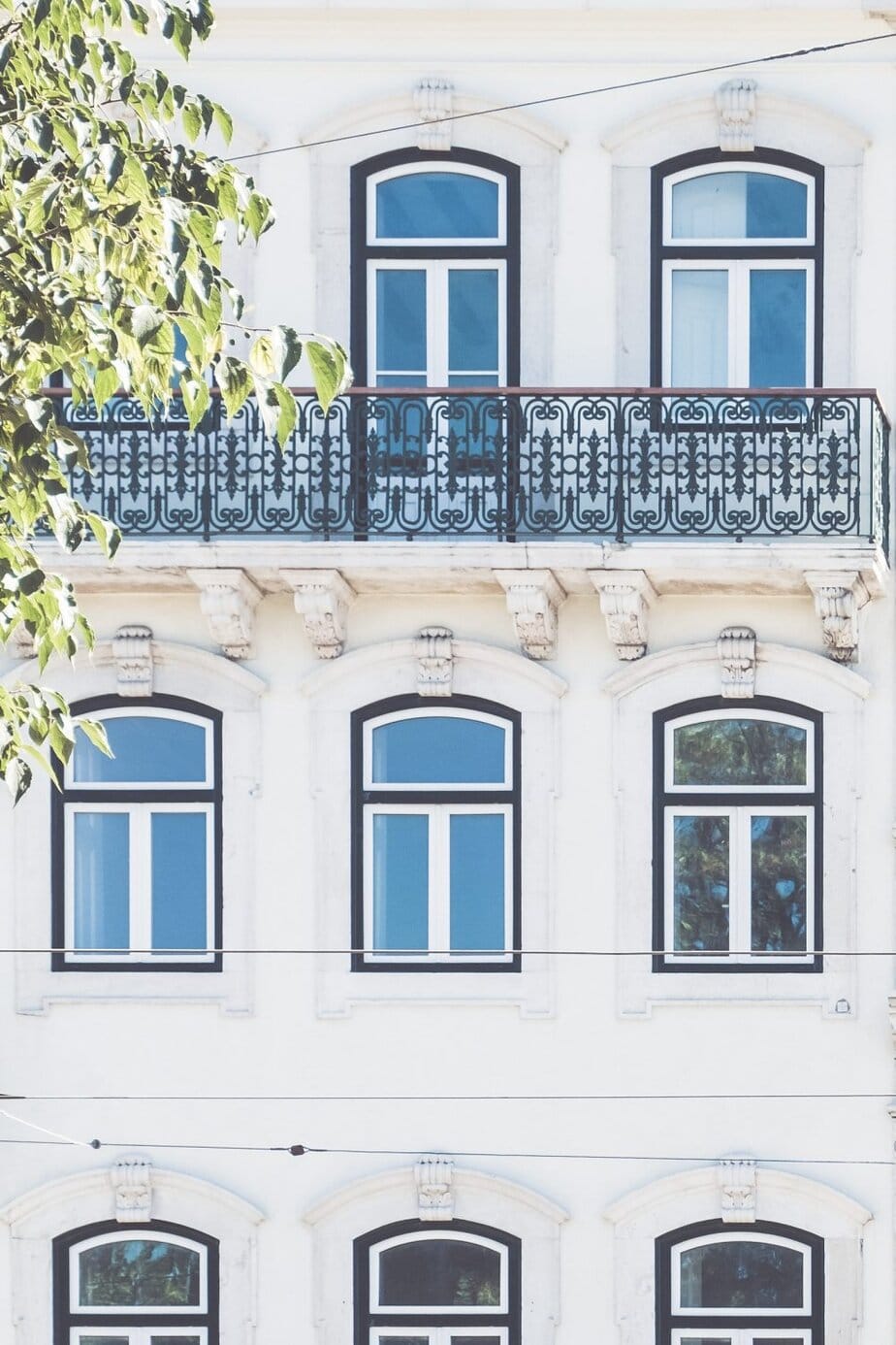 A white building with arched windows, a decorative black balcony railing, and ornate architectural details, partially obscured by tree branches on the left.