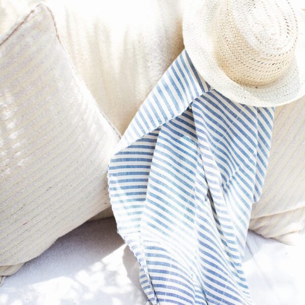 A straw hat and a blue-and-white striped scarf rest on a cream-colored cushion in bright sunlight.