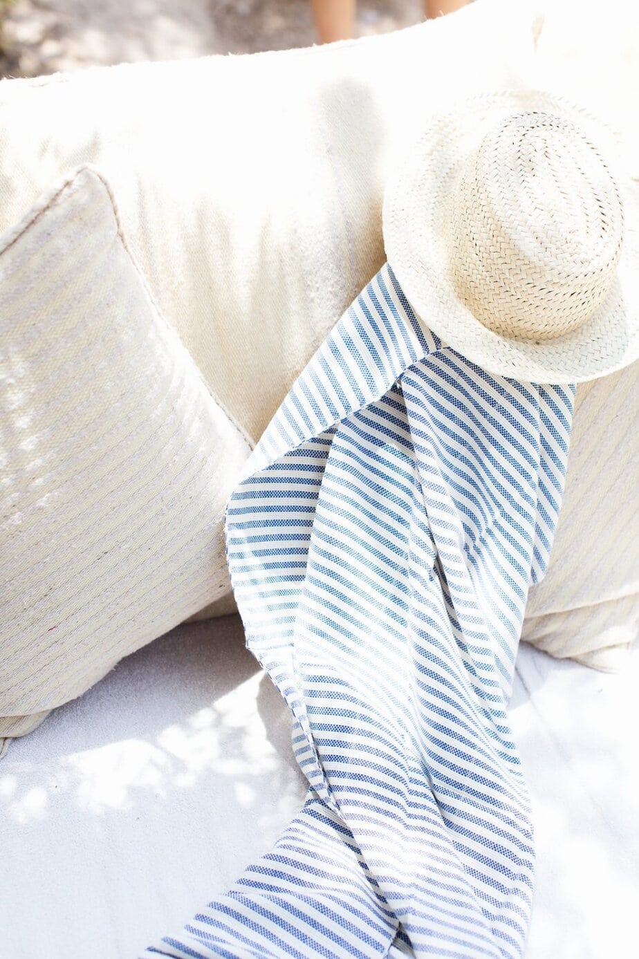 A straw hat and a blue-and-white striped scarf rest on a cream-colored cushion in bright sunlight.