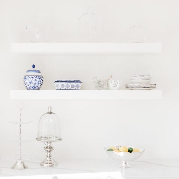 Minimalist white kitchen with floating shelves displaying blue and white ceramics, glassware, silver trays, and a bowl of lemons and limes on the counter. Minimalist white kitchen with floating shelves displaying blue and white ceramics, glassware, silver trays, and a bowl of lemons and limes on the counter.