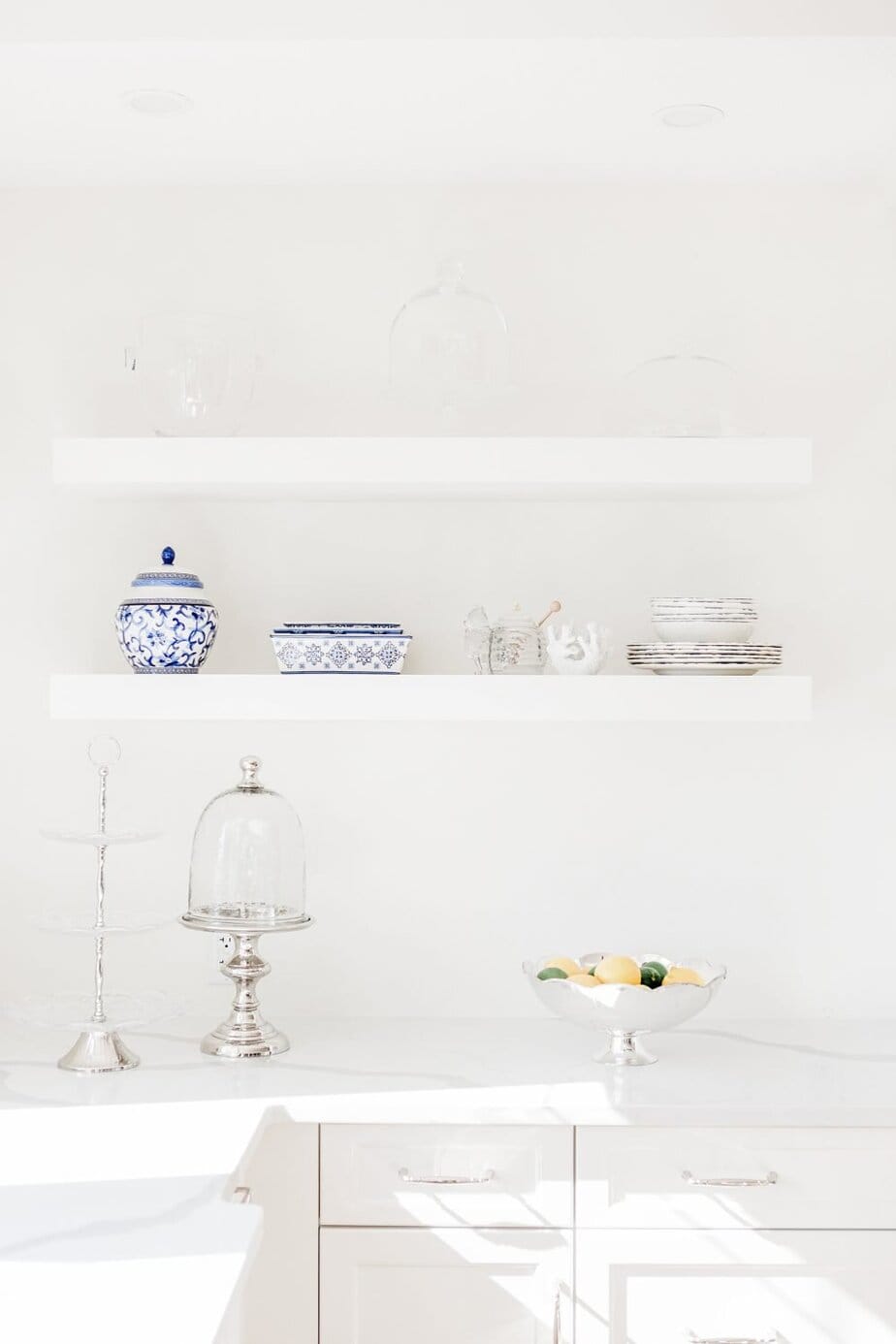Minimalist white kitchen with floating shelves displaying blue and white ceramics, glassware, silver trays, and a bowl of lemons and limes on the counter.