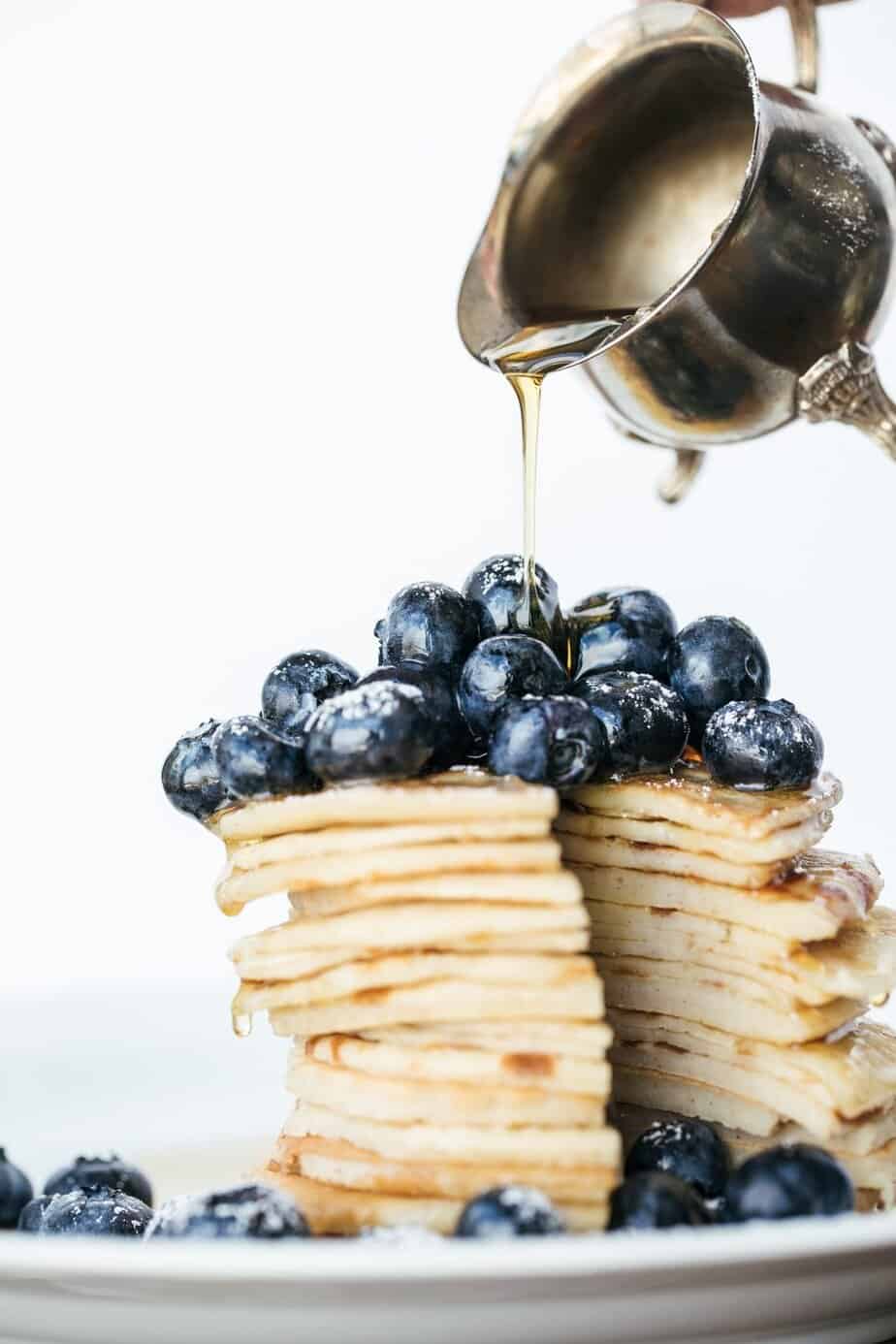 A stack of pancakes topped with blueberries is being drizzled with syrup from a silver pitcher.