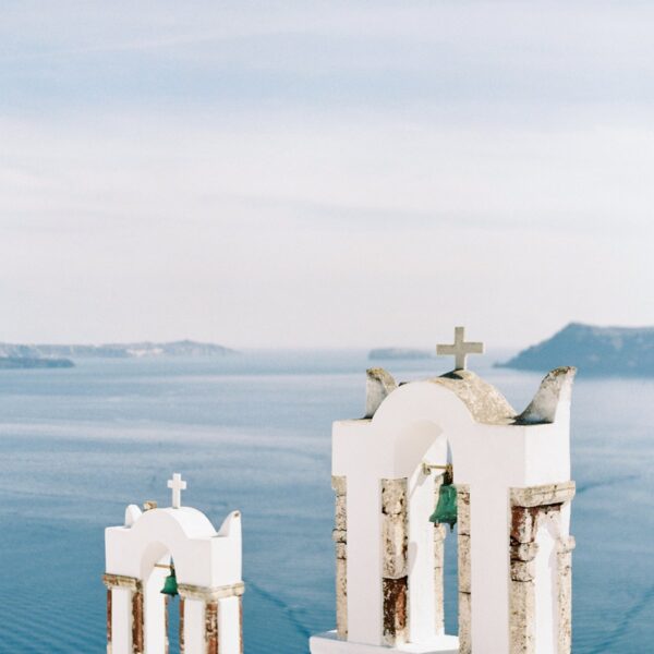 White bell towers with crosses overlook the blue sea and distant islands under a clear sky, characteristic of Santorini, Greece.