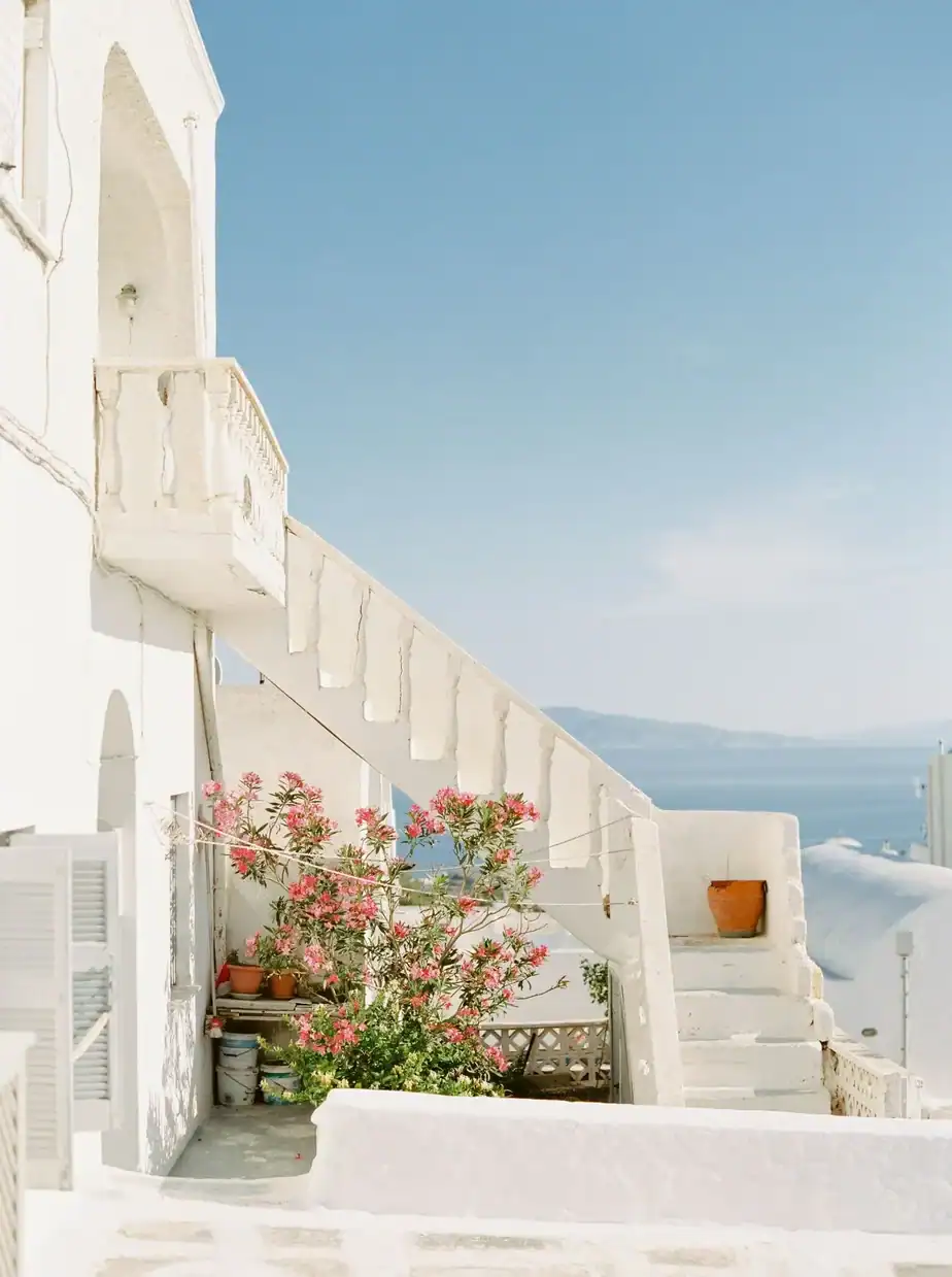 A white staircase with a potted plant and pink flowers leads up the side of a Mediterranean-style building, overlooking a clear blue sky and sea.